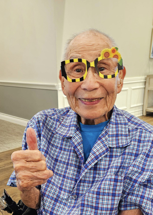 Senior resident gives a thumbs-up while wearing playful glasses during a themed event inside a senior living community.