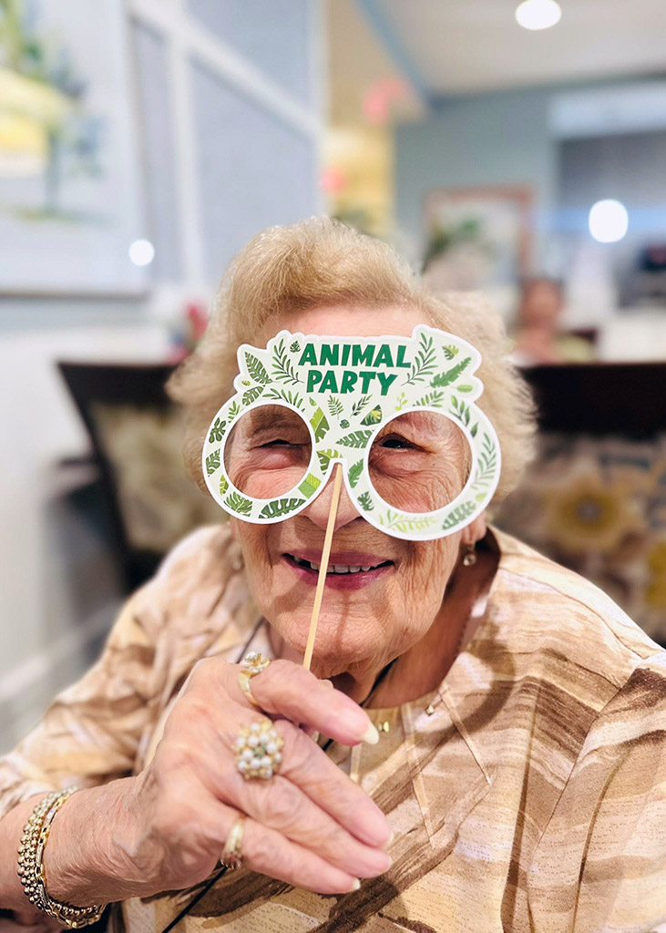 Senior resident laughs while holding playful party glasses during a themed event inside a senior living community.