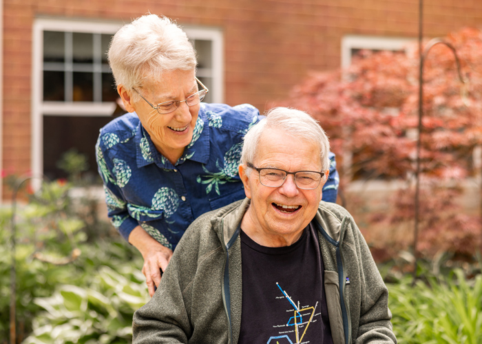 Two senior residents laugh together while spending time outdoors on a landscaped senior living community patio.